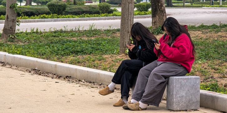 Δύο νεαρές γυναίκες κάθονται σε ένα πάρκο στο Θανχ Χόα του Βιετνάμ, στις 18 Μαρτίου 2025, συγκεντρωμένες στα smartphones τους. [MICHAEL NGUYEN/NurPhoto μέσω AFP]