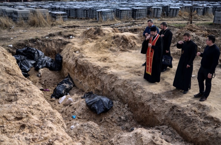 Ιερείς προσεύχονται δίπλα σε σάκους πτωμάτων σε ομαδικό τάφο που βρίσκεται στον περιβάλλοντα χώρο της εκκλησίας του Αγίου Ανδρέα στην Μπούτσα της Ουκρανίας, στις 7 Απριλίου 2022. [RONALDO SCHEMIDT/AFP]