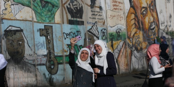 Παλαιστίνιες γυναίκες βγάζουν selfie μπροστά από τμήμα του ισραηλινού διαχωριστικού τείχους που είναι καλυμμένο με γκράφιτι και δείχνει τον φυλακισμένο ηγέτη της Φατάχ, Μαρουάν Μπαργούτι, στη Δυτική Όχθη, στις 5 Απριλίου 2014. [ABBAS MOMANI/AFP]