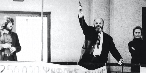 Ο Βασίλης Τσιρώνης, κραδαίνοντας το πιστόλι, στο μπαλκόνι του διαμερίσματος που είχε ανακηρύξει ως αυτόνομο «Κράτος του ΟΕΜ». Δεξιά, η γυναίκα του, Βαρβάρα, και αριστερά ο γιος του, Κώστας.