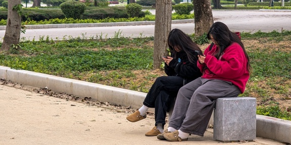 Δύο νεαρές γυναίκες κάθονται σε ένα πάρκο στο Θανχ Χόα του Βιετνάμ, στις 18 Μαρτίου 2025, συγκεντρωμένες στα smartphones τους. [MICHAEL NGUYEN/NurPhoto μέσω AFP]
